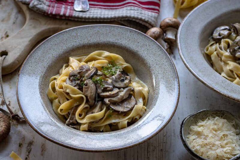 Ein Teller mit Nudel-Pilz-Pfanne in Miso Rahmsoße, zubereitet mit Champignons, Austernpilze, Shiitake-Pilzen und Parmesan, garniert mit frischen Kräutern