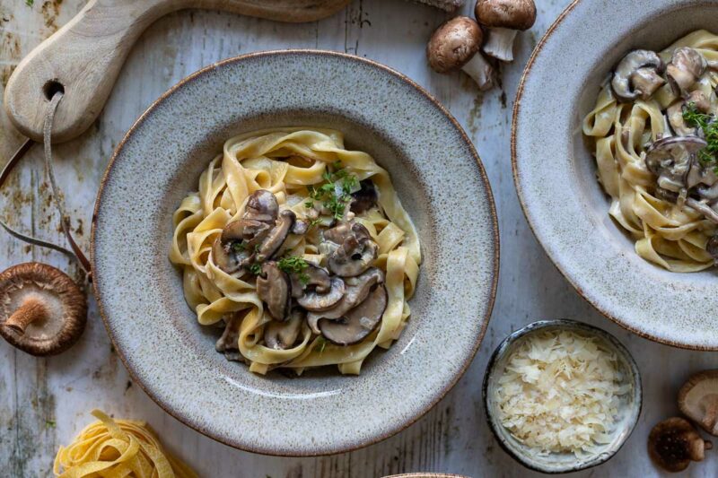 Teller mit Nudel-Pilz-Pfanne zubereitet mit Champignons, Austernpilze, Shiitake-Pilzen, Miso Rahmsoße,und Parmesan, garniert mit frischen Kräutern